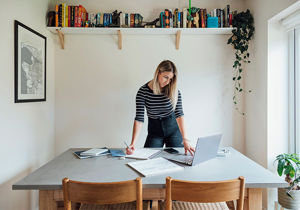 A female entrepreneur working from home to create unique jewelry designs on her laptop, she is standing with a wide smile as she looks at the camera.
